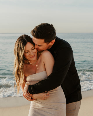 A couple embraces on a beach at Marquis Los Cabos, ocean visible behind them at sunset