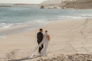 A bride and groom walk barefoot along a beach at Marquis Los Cabos, waves rolling in beside them